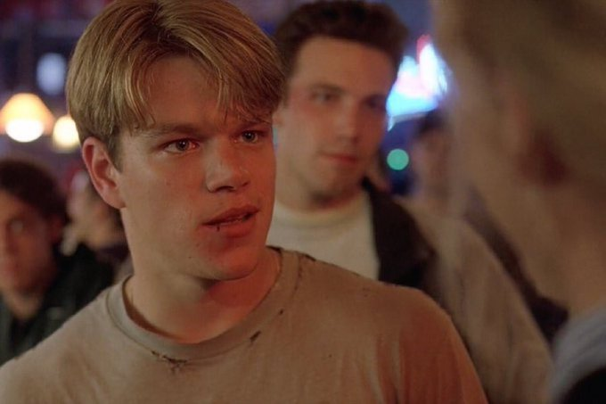 A young man with dirty blonde hair and a worn t-shirt looks intently at someone off-camera with a serious, confrontational expression, while another man stands behind him in what appears to be a dimly lit bar or social setting. The image, featuring actor Matt Damon from the film "Good Will Hunting," captures the intense debate-ready gaze of someone ready to dismantle an opposing argument.