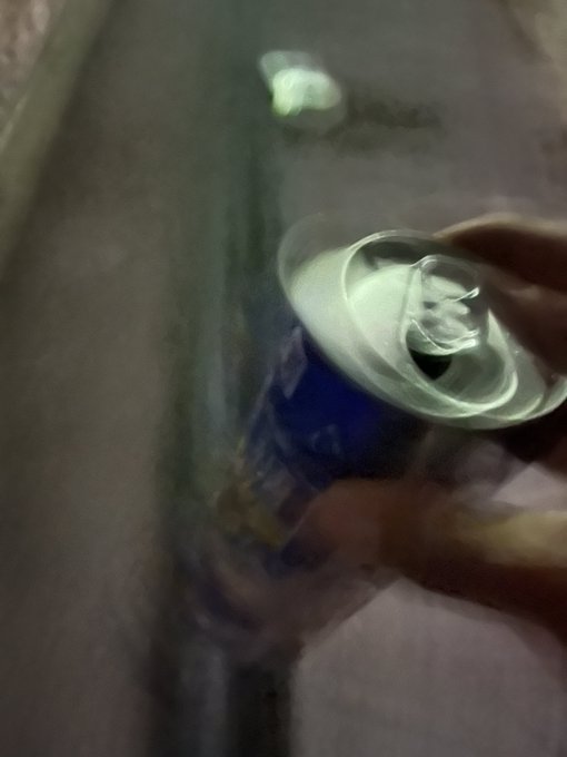 The image shows a person holding an open can of soda, possibly a root beer or another type of soft drink. The background appears blurry, but there are two other people visible in the scene. The main focus of the photo seems to be on the soda can and its contents rather than the surrounding environment.