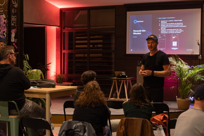 The image shows a man standing at a podium, giving a presentation or speech about Bitcoin basics. The audience consists of several people sitting in chairs around the room, attentively listening to the speaker. A large screen displaying information related to Bitcoin can be seen behind the speaker and the audience. In addition to the main speaker, there are other individuals present who may also have an interest in Bitcoin or blockchain technology.