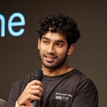 A man with curly hair and a beard speaks into a microphone while wearing a black "Movement Labs" t-shirt with Japanese text 流動ラボ.