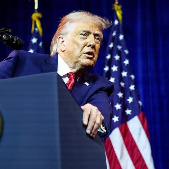 Donald Trump, in a dark suit with a red tie, standing at a podium and gesturing with his arm against a backdrop of American flags and dark blue curtains.