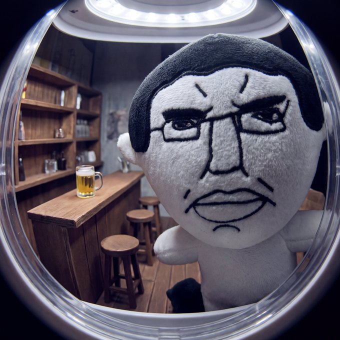 A plush doll with an angry, drawn-on face is shown through a fisheye lens in a bar setting, with a mug of beer visible on the counter behind it.