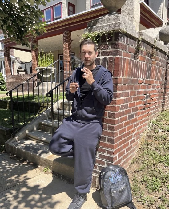 A man in a dark blue tracksuit standing on steps beside a brick wall, holding a smoking cigarette, with a Louis Vuitton checkered backpack at his feet.