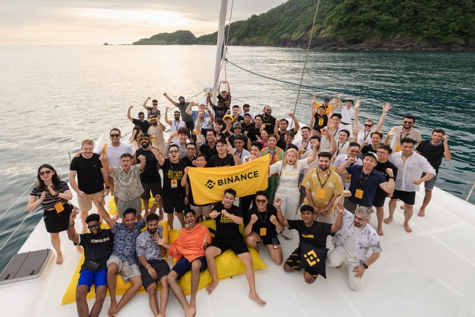 A group of people on a sailboat hold up a yellow "BINANCE" flag and give thumbs-up gestures to the camera. This lively gathering of Binance team members captures the energetic atmosphere of Day 1 of the Binance Campus Asia event out on the water.