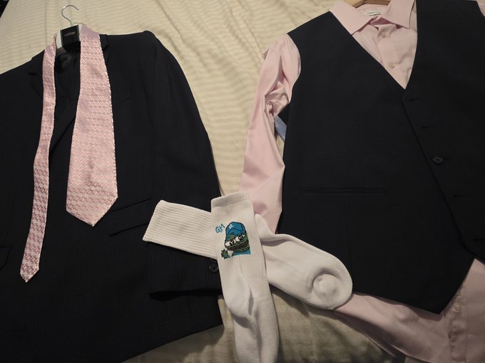 Two formal outfit combinations laid out on a bed, each consisting of a black vest with either a black jacket and pink tie or a pink button-up shirt, alongside white socks featuring a cartoon character design.