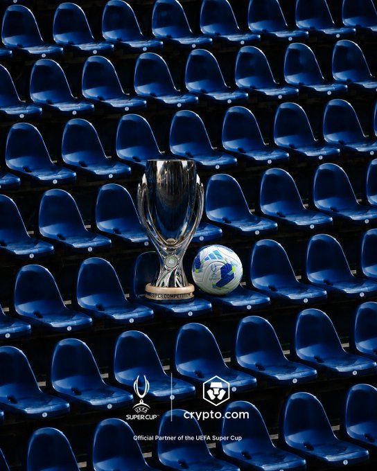 The UEFA Super Cup trophy sits atop blue stadium seats beside an official match ball, with a "crypto.com" logo and "Official Partner of the UEFA Super Cup" text displayed at the bottom, symbolizing the anticipation and grandeur of the upcoming championship match.