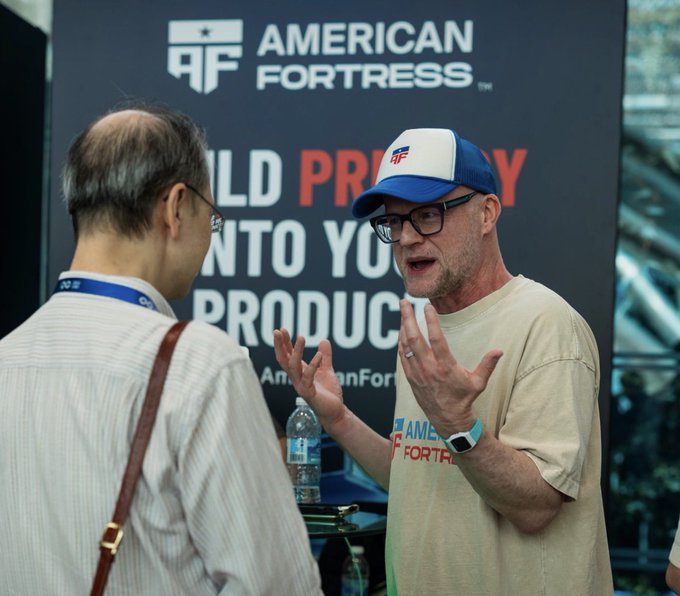 Two men converse at what appears to be an American Fortress event, with one wearing a tan t-shirt and American Fortress cap speaking animatedly to a man in a striped shirt and lanyard.