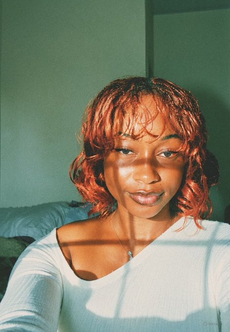 A young woman with wet, reddish-orange hair and a serious, unsmiling expression looks directly at the camera, wearing a white long-sleeve top and a delicate necklace, her face dramatically lit by a window casting warm light. Her deliberate frown and the post's caption reference "Day 1 of not smiling," perfectly matching the stoic, almost sullen mood of her portrait as she waits for a stock mentioned ($ANSEM) to multiply.