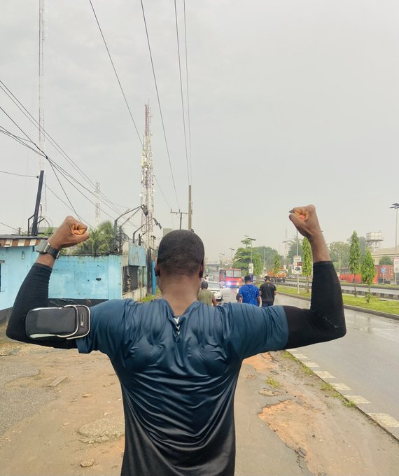 A person in a blue and black athletic shirt stands on a roadside with their back to the camera, flexing both biceps in a power pose.