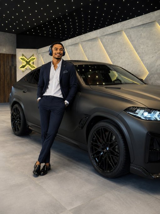 A man in a navy blue suit with headphones stands next to a matte black BMW SUV in a showroom with an LED star ceiling and neon X logo on the wall.