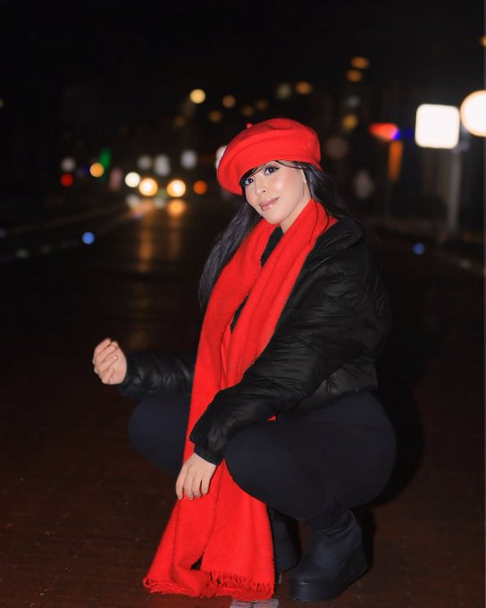 A woman wearing a bright red beret, a matching red scarf, and a black puffer jacket crouches on a wet city street at night with blurred traffic lights in the background.