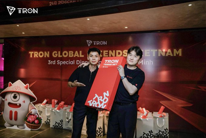 Two men pose at a "TRON GLOBAL FRIENDS VIETNAM" 3rd Special Dinner event, holding red "TRON 1997" gift bags with a mascot and event gifts displayed below.