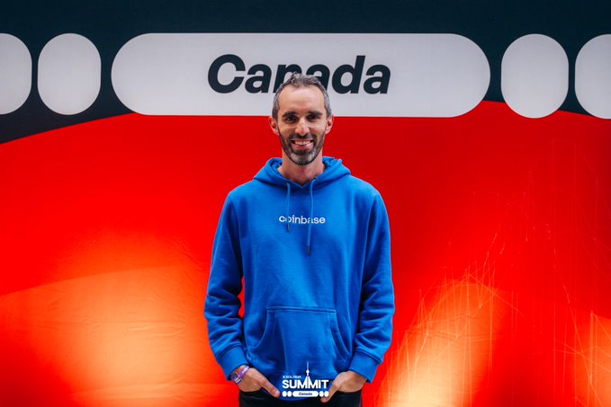 A man wearing a blue 'coinbase' hoodie stands smiling in front of a large red screen displaying the word 'Canada' and the text 'SUMMIT'.