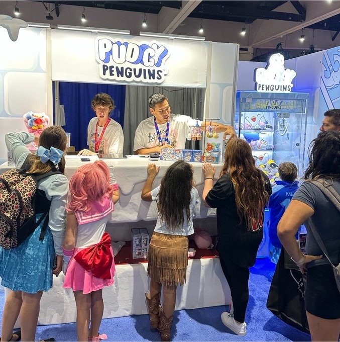 Children in colorful costumes gather at a "Pudgy Penguins" convention booth, where brand representatives engage with fans and showcase merchandise, illustrating the community-driven appeal the post is inviting content creators to join.