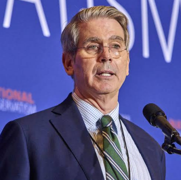 A man in glasses, a blue suit, and a striped green tie speaks into a microphone at a podium.