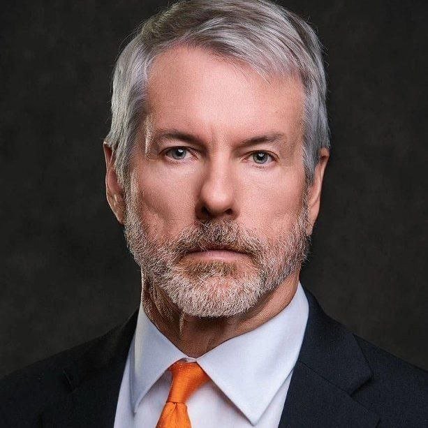 A professional studio portrait of a middle-aged man with gray hair and a beard, wearing a dark suit, light blue shirt, and orange tie, against a dark gray background.