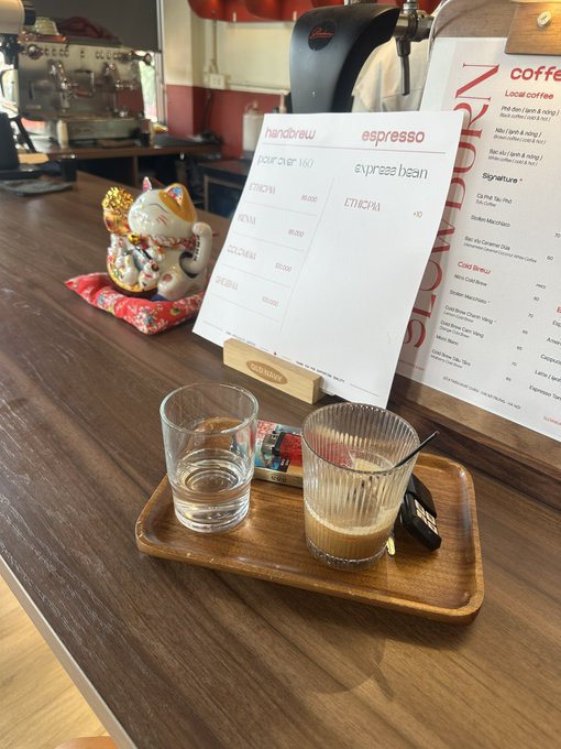 The image shows a wooden table with two glasses of coffee, one filled with water and the other with milk. There are also some papers or menus on the table that appear to be related to the coffee shop's offerings. The text "Gm cả nhà mình Ngày mới vv nhé" is present in the image, which could possibly be a description of the coffee shop's specialty or a menu item.