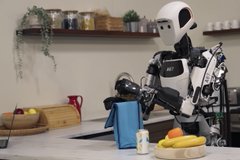 A humanoid robot in a kitchen environment holding a blue cloth near a counter with a bowl of fruit, appearing to assist with household tasks.