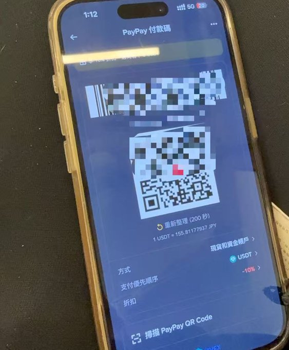 A smartphone displays a PayPay payment screen with a Japanese text header 'PayPay 付款碼' and a central QR code, showing an exchange rate of '1 USD ≈ 155.8177937 JPY' over a dark background.