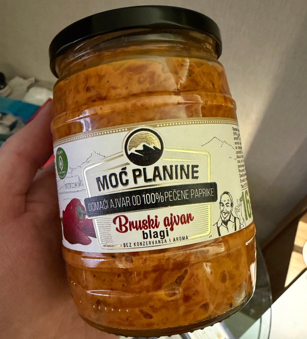 Hand holding a glass jar of 'MOĆ PLANINE' brand ajvar, a traditional Balkan red pepper spread, labeled 'Bruski ajvar blagi' (mild), made from 100% roasted peppers, without added preservatives or aromas.