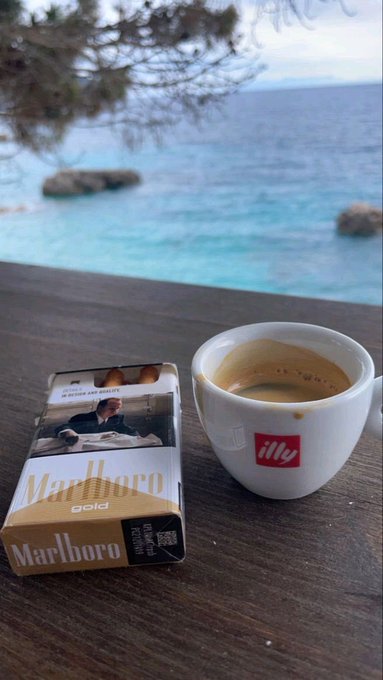 A pack of Marlboro Gold cigarettes and a cup of espresso in an 'illy' branded cup sit on a wooden table overlooking a blue ocean.