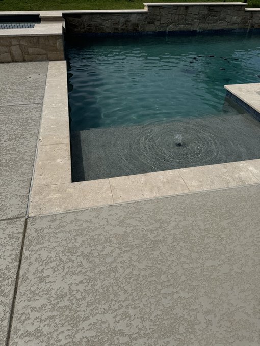 A backyard swimming pool with a stone waterfall feature is shown, featuring a shallow sun shelf with a circular drain cover and textured concrete decking.
