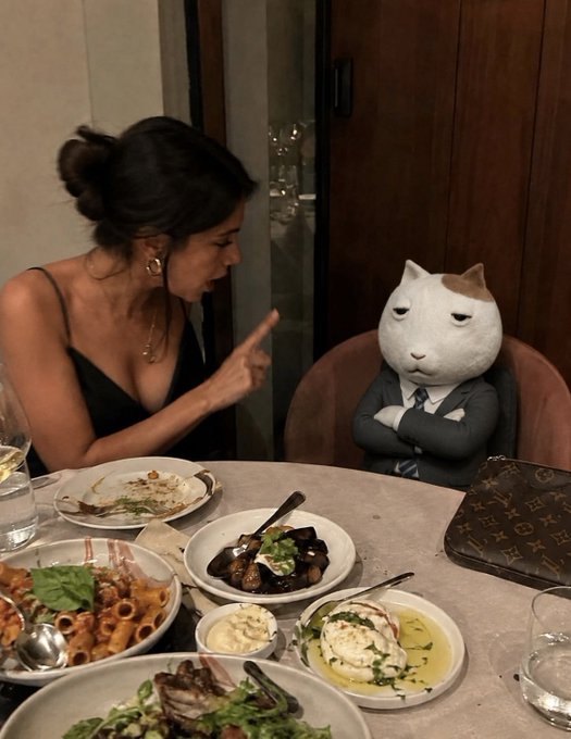 A woman in a black dress sits at a restaurant table with various dishes, pointing her finger at a person wearing a large cartoon cat head and a business suit.