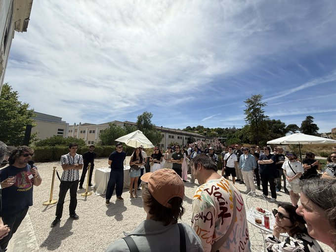 The image shows a group of people gathered outdoors, possibly at an event or gathering. They are standing around each other, engaged in conversation and enjoying their time together. The sky above them appears cloudy, suggesting that they might be experiencing some weather conditions during this outdoor social event. In the background, there is a building visible, which could be part of the venue for the gathering.