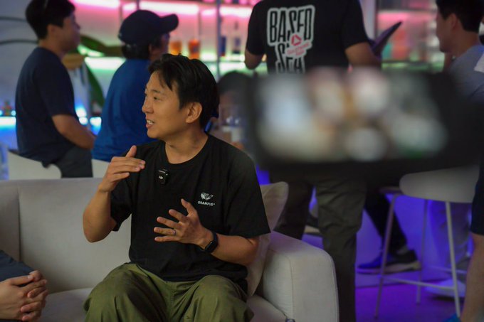 A man sits on a white couch gesturing with his hands while engaged in conversation, wearing a black t-shirt and olive green pants, with other people visible in the background of what appears to be a social event or networking gathering.