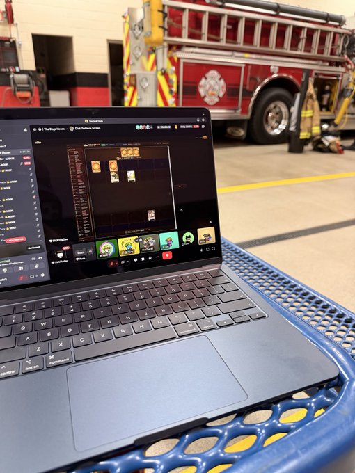 A laptop sits on a blue mesh chair, displaying what appears to be a Pixel Press game interface, with a red fire engine parked in the background of the fire station.