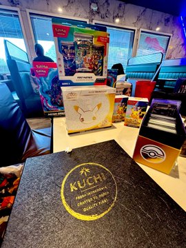 A table at a KUCHI restaurant displaying a branded menu with the gold 'KUCHI' logo, alongside boxes of branded merchandise including earbuds and figurines.