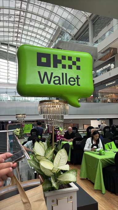 A large green OKX Wallet promotional balloon display floats above a table setup with green tablecloths in a shopping mall atrium, likely for a crypto event.