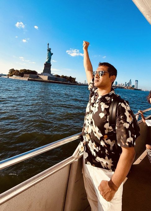 A man in a patterned shirt and sunglasses poses with a raised fist on a boat, with the Statue of Liberty visible in the background against a blue sky.