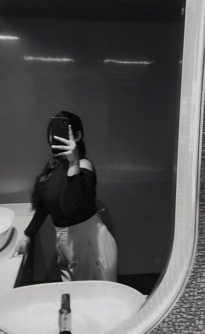 A black and white mirror selfie of a woman wearing an off-shoulder top and light-colored skirt/pants, holding a phone to take the photo.