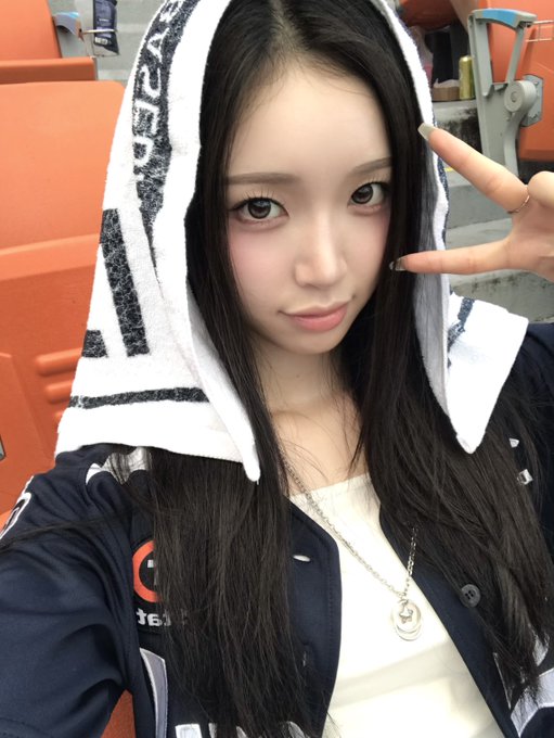 A close-up selfie of a young woman with long dark hair, wearing a navy jacket and a towel with text draped over her head, making a peace sign with her long nails.