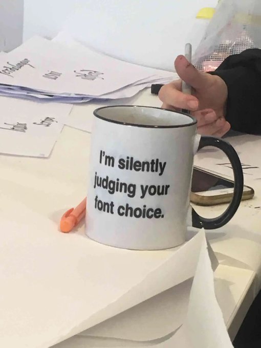 A white ceramic mug with black rim and handle sits on a desk, displaying the humorous text 'I'm silently judging your font choice,' with a hand visible holding a pen in the background.