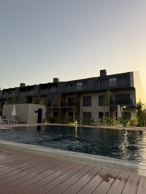 A modern, dark-colored multi-story building stands behind a swimming pool, with wooden decking in the foreground and lounge chairs arranged poolside under a clear evening sky.
