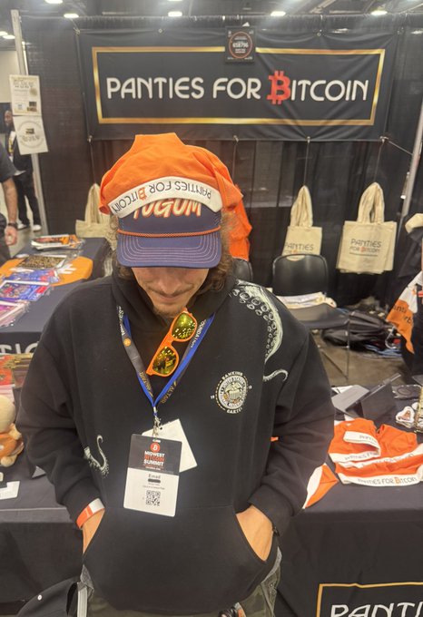 A person wearing an orange 'PANTIES FOR BITCOIN' hat and black hoodie stands at a convention booth with the same branding on the banner behind them.