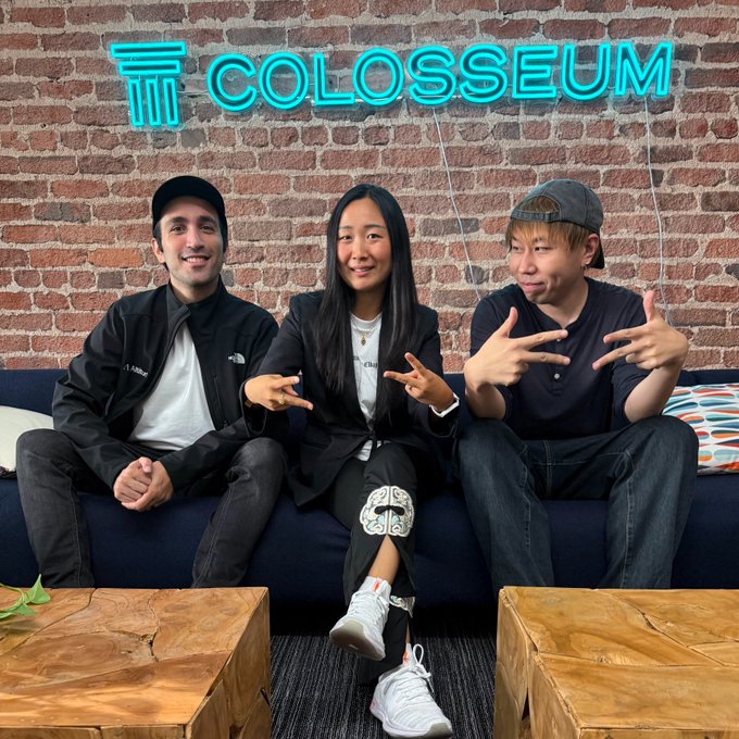 Three people sitting on a blue couch in front of a brick wall with a 'COLOSSEUM' neon sign, all making hand gestures toward the camera.