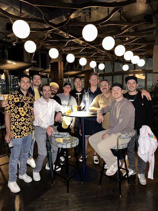 A group photo of nine men smiling and posing together at a venue decorated with hanging round lights and twisted branches on the ceiling.