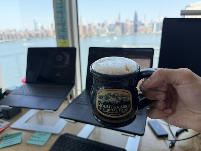 A hand holds up a dark glass mug filled with a frothy beverage in front of two open laptops on a desk; the mug features a 'Mount Rainier National Park Established 1899' logo, with a sunny city skyline visible across water through the window behind.