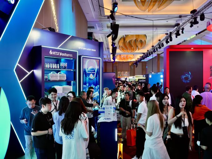 A crowded exhibition floor with people networking around bright, colorful booths, prominently featuring "HTX Ventures" branding in a blue-lit display area.