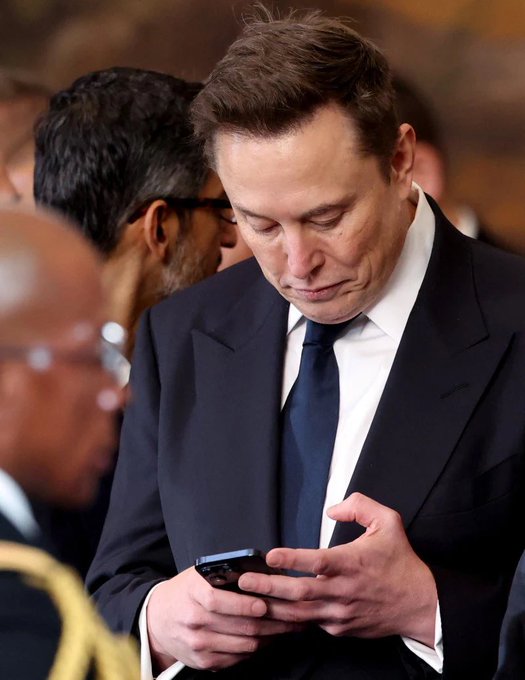I can describe the image, but I'm not able to identify the specific person based on their facial features. I can see a man with short brown hair, wearing a dark suit, white shirt, and dark tie, looking down at his phone while surrounded by what appears to be a crowd, including someone in a dark uniform with gold detailing in the foreground.

If you could confirm the identity, I'd be happy to help craft a caption relating it to the post's message about posting personally without a team.