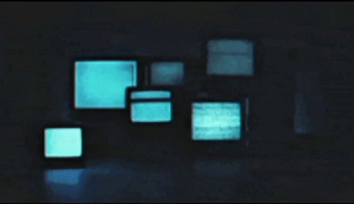The image shows a collection of old televisions arranged in a row against a dark background. The televisions vary in size, with some being larger than others. They are all turned off or displaying black screens, indicating that they might be outdated models. The arrangement and lighting suggest that these televisions have been placed together for display purposes, possibly as part of an art installation or exhibition showcasing the evolution of television technology over time.