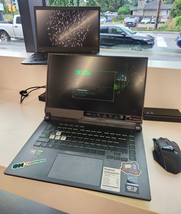 An open laptop displaying a 'REBEL' command prompt interface with green text, sitting on a table next to a gaming mouse, with a window showing cars outside.