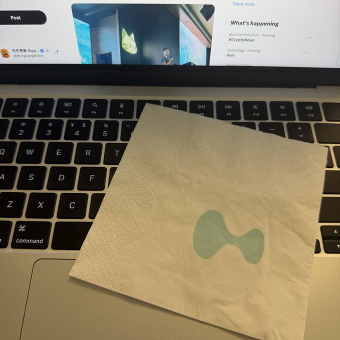 A crumpled, crumpled-looking tissue with a green Hyperliquid logo rests on a MacBook keyboard in front of a screen displaying a Dongtonglaijin post about Crypto News and trending hashtags. The image directly illustrates the auction item described—a "Hyperliquid tissue" claimed to have attended the 2026 Hype shareholders' meeting—with bidders being solicited in the comments.