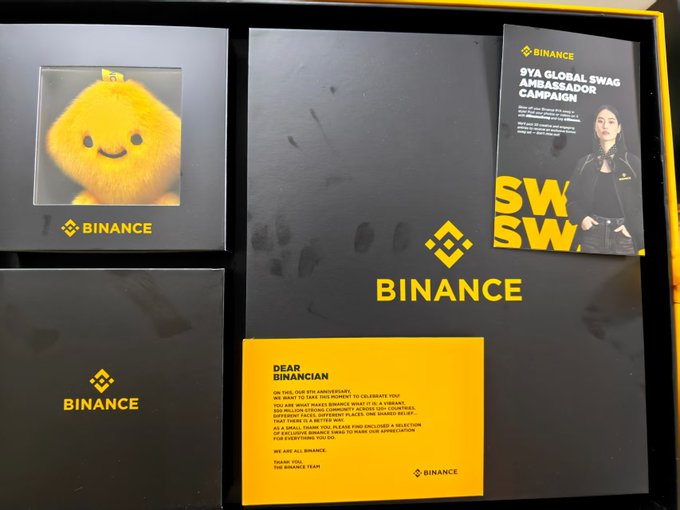 The image shows a Binance 9th anniversary swag box featuring a plush yellow mascot, branded merchandise with the "BINANCE" logo, and a card for the "SYA GLOBAL SWAG AMBASSADOR CAMPAIGN," alongside a thank-you letter from Binance addressed to "DEAR BINANCIAN" celebrating the milestone. This corresponds to the post highlighting the user's excitement about receiving Binance's 9th-anniversary merchandise, their long history of receiving yearly gifts from Binance since its 3rd anniversary, and well-wishes for the exchange's birthday.