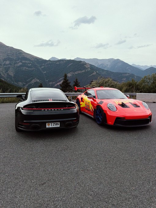 Two Porsche 911 sports cars—one sleek black and one vibrant red with racing stripes—pose back-to-back on a mountain overlook, embodying the "Lightning" theme with their striking contrast and bold presence against the alpine backdrop. The black Porsche displays a Swiss license plate reading "S 2506."