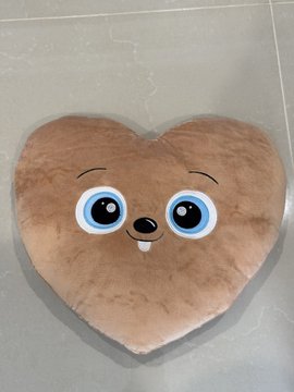 A heart-shaped brown plush cushion with cartoon eyes and a small tooth, placed on a gray floor.