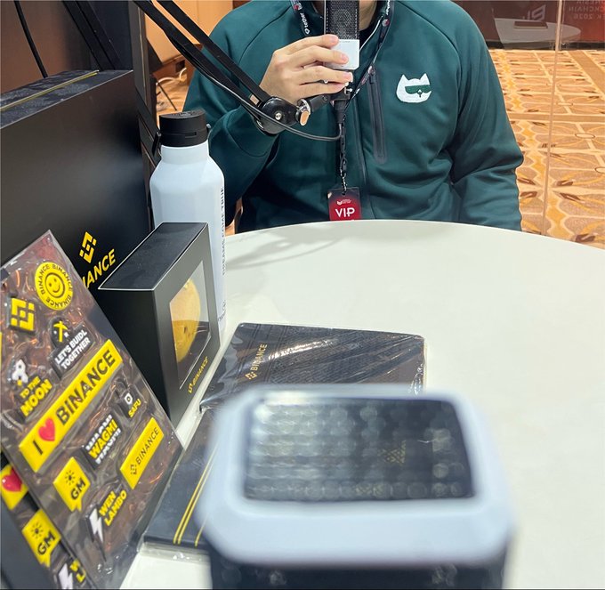 A person wearing a green sweatshirt with a Cat logo and a VIP lanyard holding a phone on a selfie stick, sitting at a table with Binance-branded merchandise including stickers, a water bottle, and boxes.
